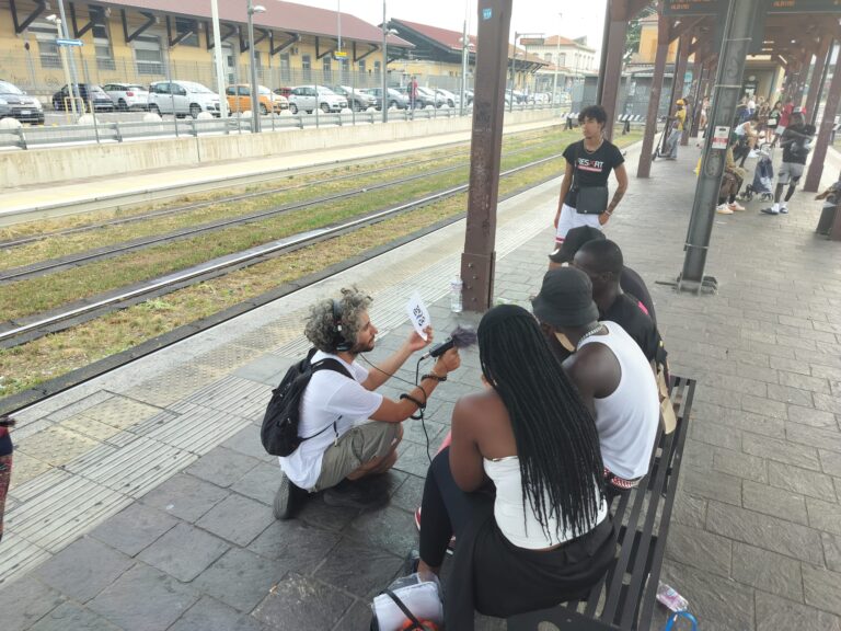 Tre ragazzi e una ragazza siedono su una panchina alla fermata della TEB in stazione. Un altro ragazzo e in ginocchio davanti a loro e tende un microfono verso uno dei ragazzi, registrando ciò che sta dicendo.