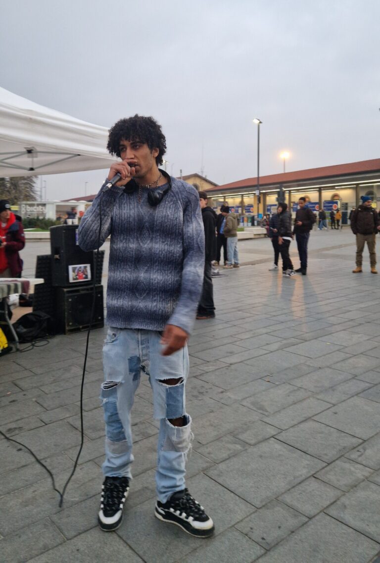 Un ragazzo canta al microfono nel piazzale della stazione. è alto, ha i capelli ricci e porta delle cuffie bluetooth appese al collo.