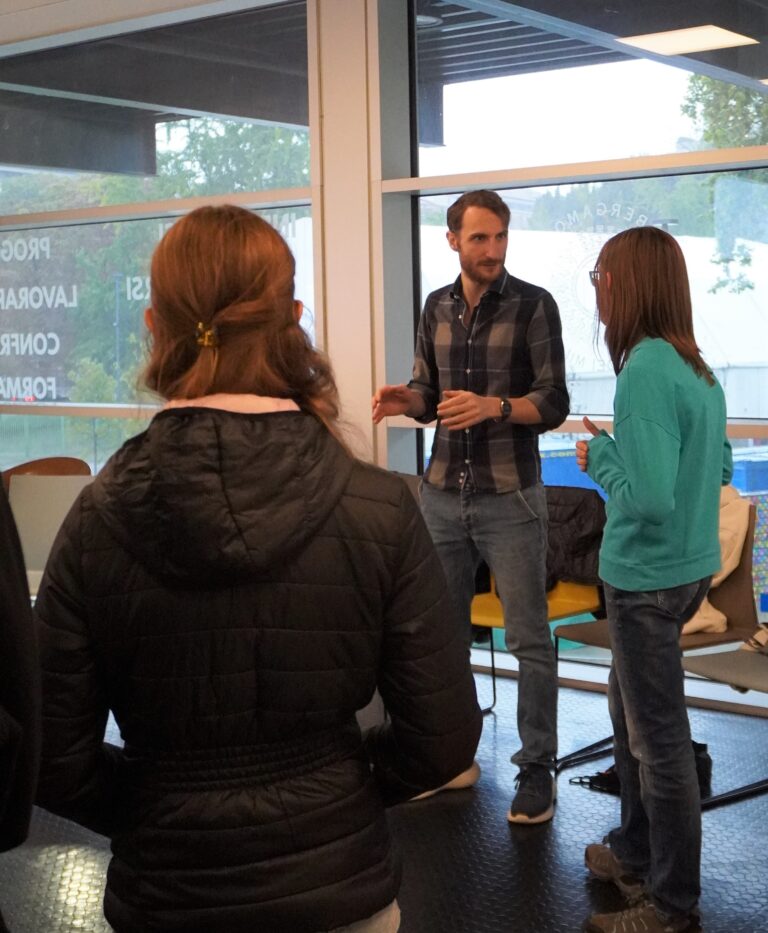 Un gruppo di studenti di terza durante un incontro di orientamento. Il gruppo è ripreso di spalle, mentre un consulente di orientamento parla. Il consulente di orientamento ha la barba, i capelli corti e una camicia.