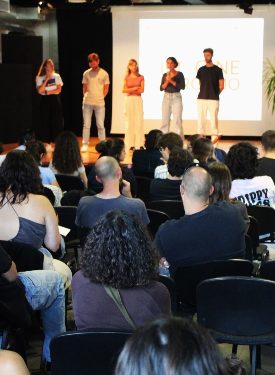 Tre ragazze e due ragazzi sono sopra un palco. Una delle ragazze sta parlando al microfono, rivolta a un pubblico fotografato di spalle.
