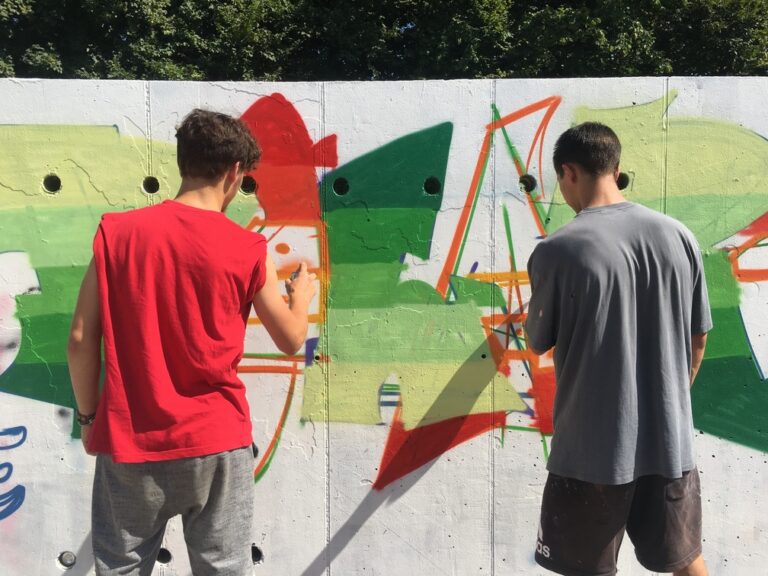 Due ragazzi sono in piedi davanti a un muro bianco su cui stanno realizzando un murales, che raffigura la scritta 