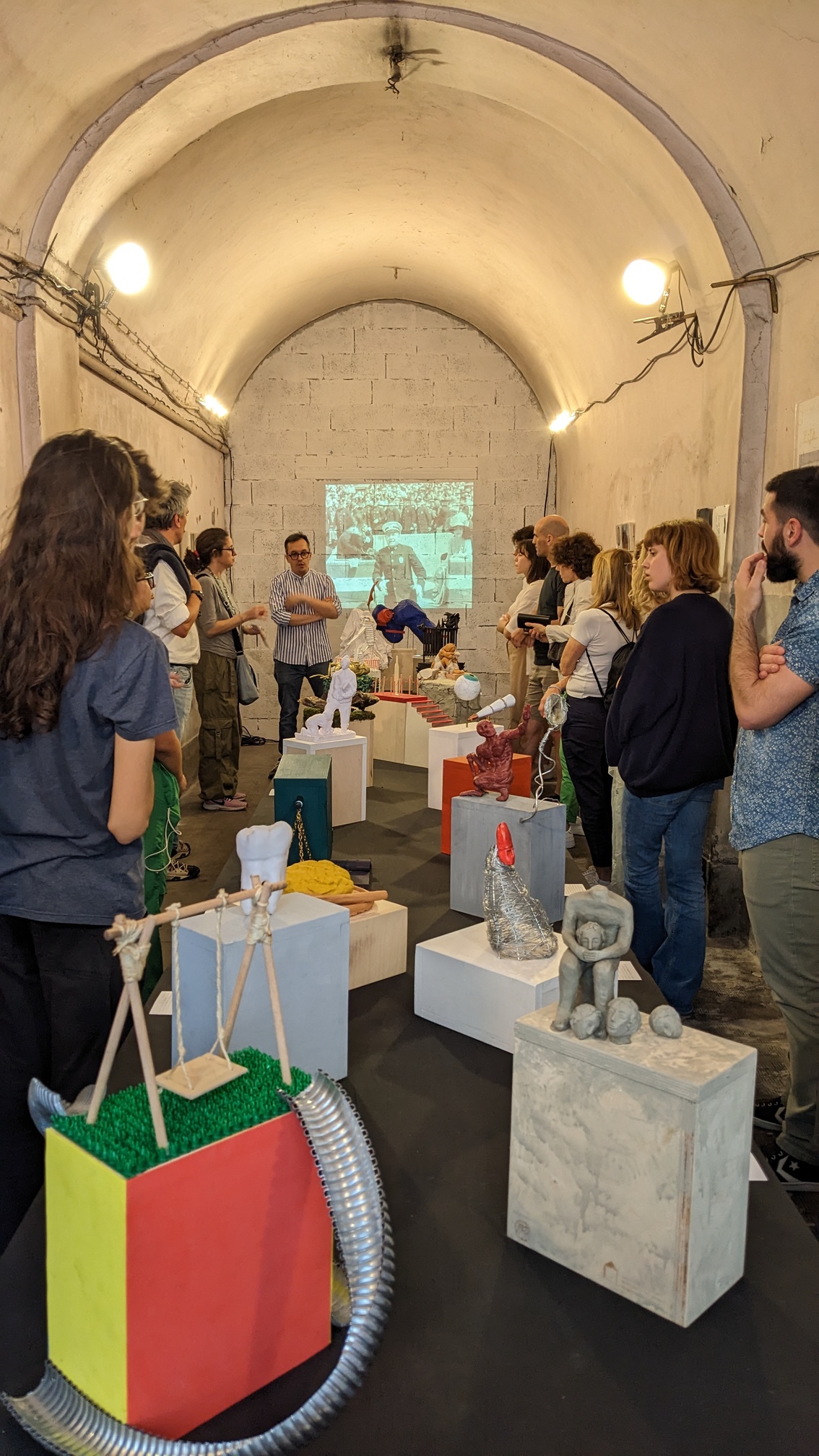 Una calsse in visita a una mostra d'arte, allestita in una stanza con soffitto a volta. La classe sta ascoltando il professore presentare le opere, principalmente sculture, che sono esposte su dei piedistalli al centro della stanza.