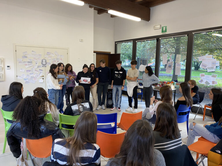 Un gruppo di studenti sta presentando al resto della classe lo sketch del loro del loro progetto. Il logo è disegnato su una foglio di carta che una ragazza tiene in mano.
