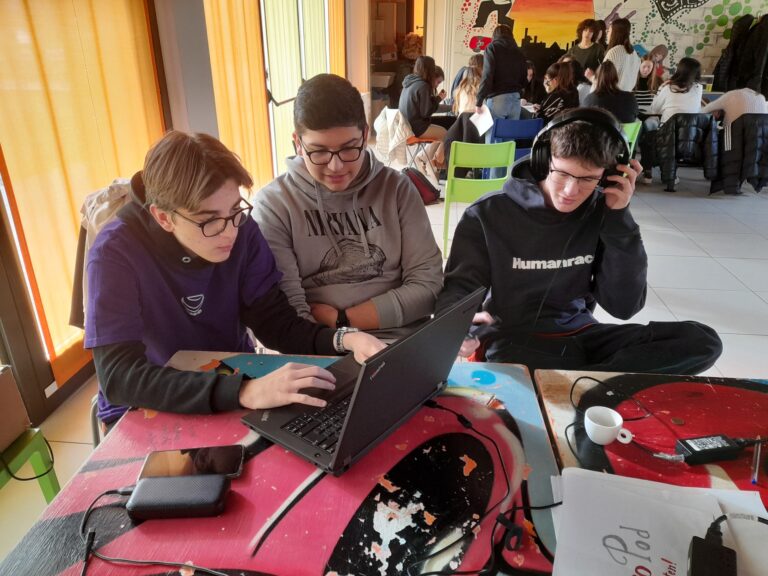 Tre ragazzi sono davanti a un pc. Uno di loro ha le mani sulla tastiera, un altro indossa grandi cuffie, il terzo osserva lo schermo.