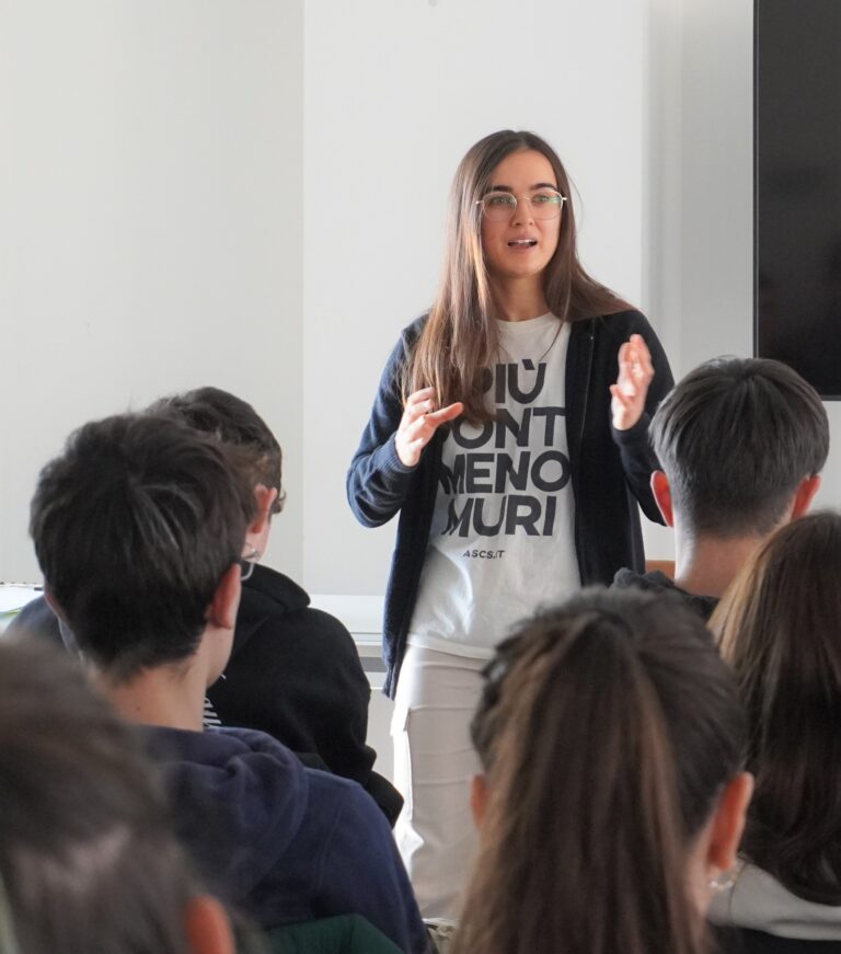 L'immagine ritrae una giovane in piedi che parla a un gruppo di studenti. Il gruppo è ripreso di spalle. La giovane indossa una t-shirt con scritte e un golf nero, ha gli occhiali e i capelli lunghi.