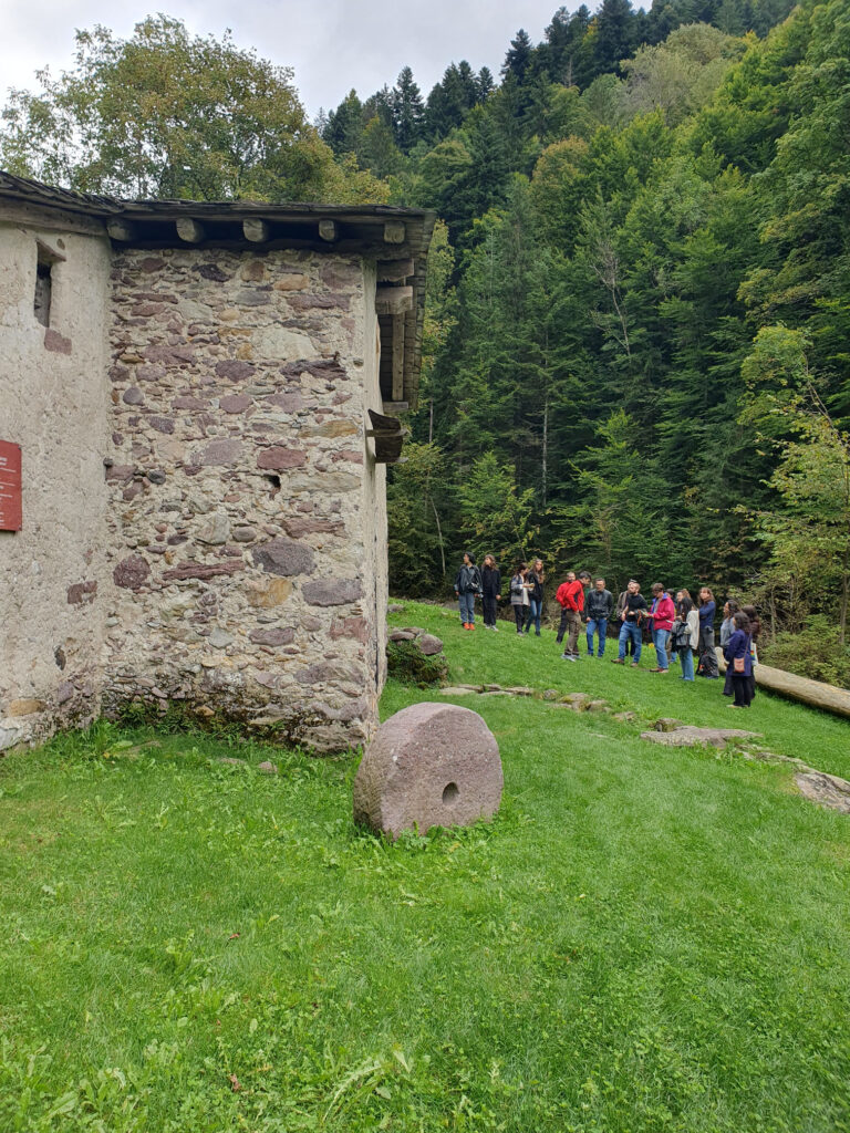 Un gruppo di persone in abiti, fotografato in lontananza, sosta vicino a una costruzione in pietra. Sono nei pressi di un bosco, in montagna.