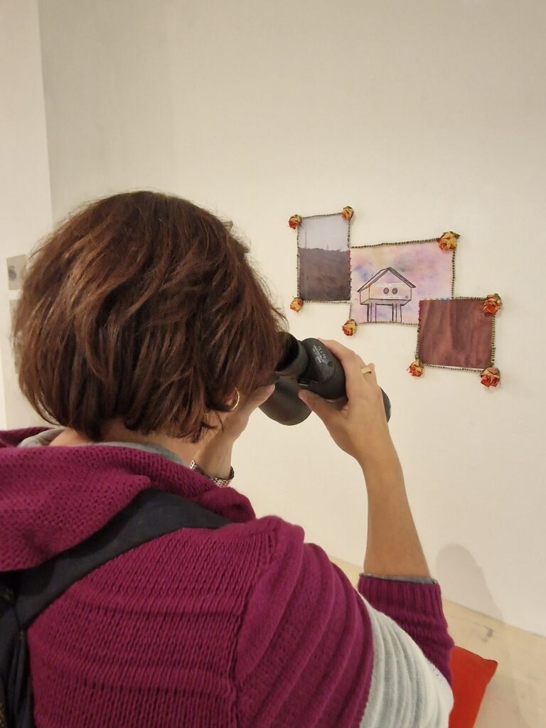 Una donna, fotografata di spalle, osserva un'opera in mostra utilizzando un binocolo, parte dell'opera stessa.