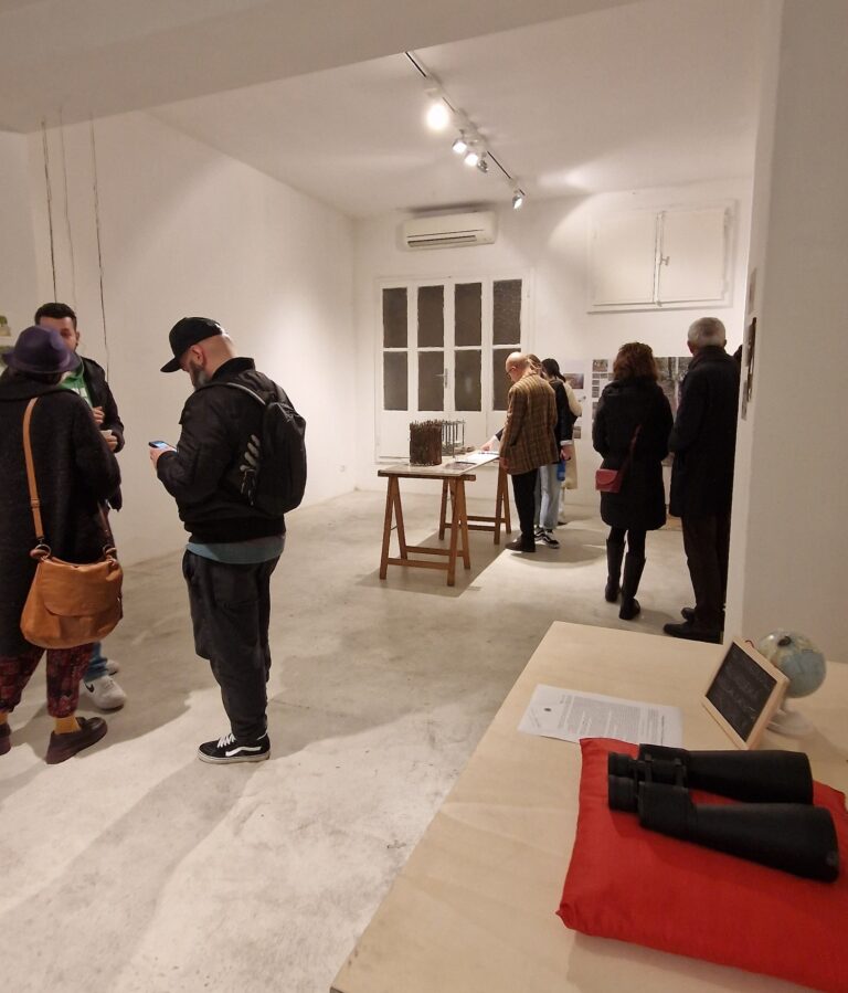 Un gruppo di persone visita una mostra d'arte, allestita. Le opere sono esposte su dei tavoli e appese alle pareti.