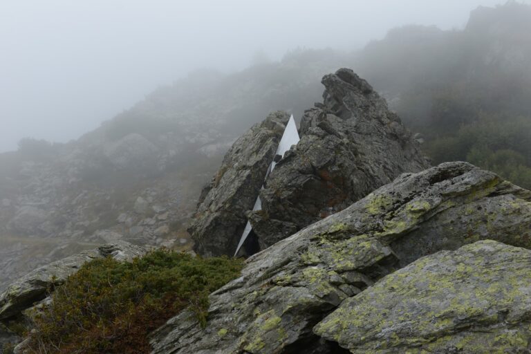 L'opera è costituita da una lastra di vetro riflettente, di forma triangolare, incastrata tra le rocce di una montagna. La foto è stata scattata in una giornata nebbiosa.