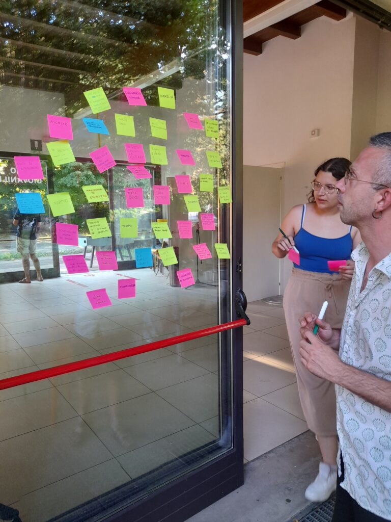 Due persone guardano una porta a vetri piena di post-it colorati. Una delle due due sta per attaccare un nuovo post-it
