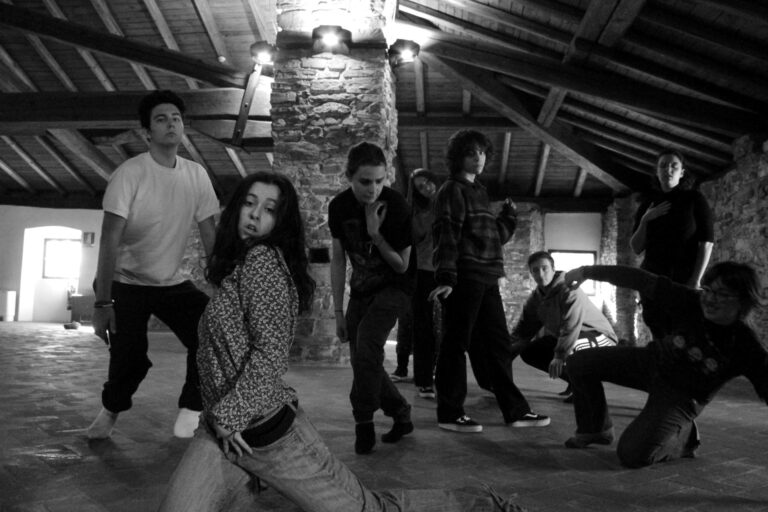 Un gruppo di otto giovani posa in una foto in bianco e nero. Ogni persona assume una posa diversa, in movimento. L'ambientazione è al chiuso, in una stanza con un soffitto di legno e pareti in pietra.