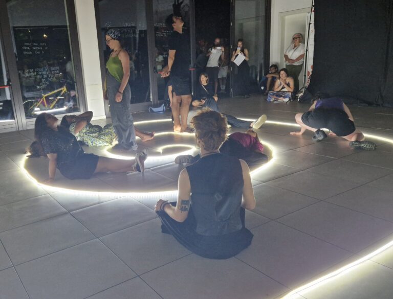 La foto ritrae il momento di una performance: un gruppo di otto giovani è distribuito all'interno di uan spirale luminosa costruita con un sistema di luce al led sul pavimento. Sono in posizione diverse: un ragazzo in primo piano siede a gambe incrociate, rivolto verso il centro della spirale; una ragazza e un ragazzo sono in piedi, altre ragazze sono sedute o distese.