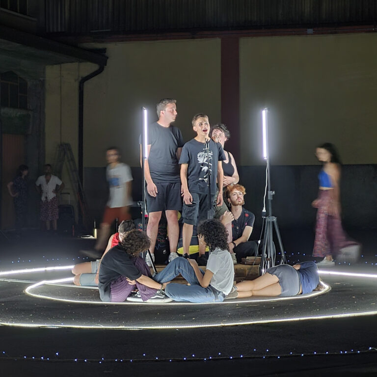 La foto ritrae un momento di una performance: un gruppo di una decina di giovani è raccolto al centro di una spirale luminosa, costruita grazie a un sistema di luci al led sul pavimento. Una parte di loro è in piedi, una parte è distesa o seduta per terra, una parte sta camminando intorno alla spirale.