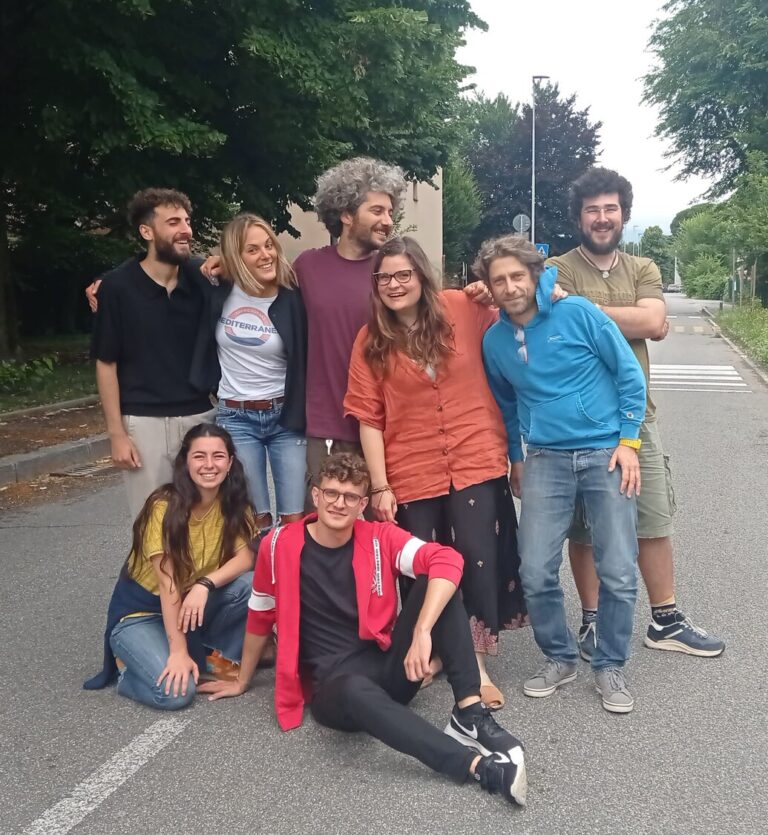 Un gruppo di persone posa abbracciato in mezzo a una strada. Due sono sedute, sei in piedi. Sono le educatrici ed educatori di Giovani Onde.