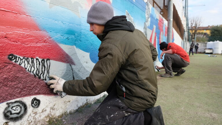 Due ragazzi sono inginocchiati vicino a un muretto e realizzano gli ultimi ritocchi di una murales con una bomboletta spray.