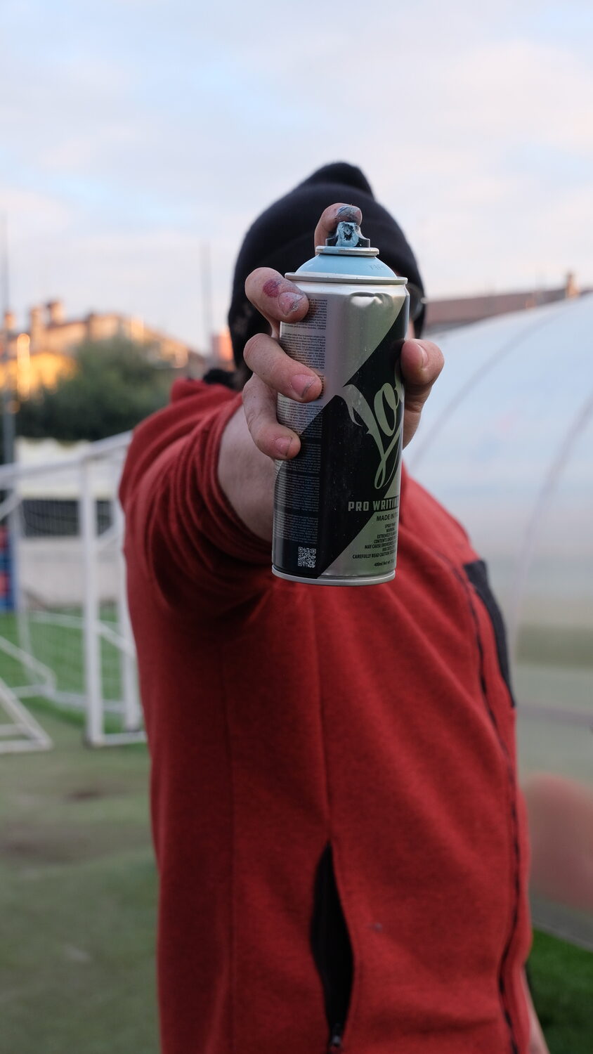 Un ragazzo punta una bomboletta spray verso l'obiettivo. La bomboletta è all'altezza del suo viso e lo copre completamente.