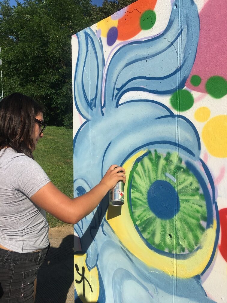 Una ragazza sta tracciando con una bomboletta spray il contorno dell'occhio gigante di un ciclope, soggetto del murales.