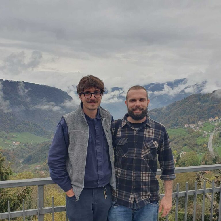 Due ragazzi vestiti con abiti sportivi da montagna sono in posa davanti a una balaustra che si affaccia su una valle. Sorridono e guardano in camera.