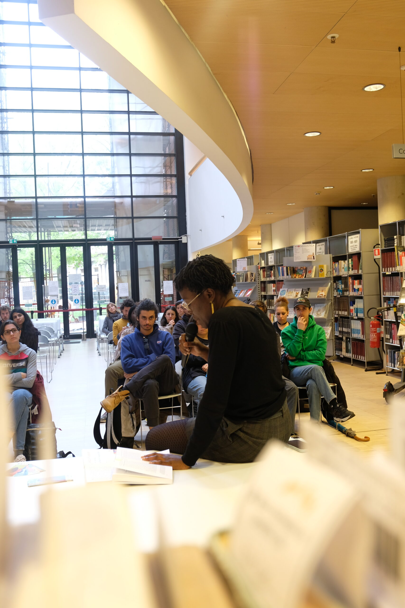 Una ragazza è seduta su un tavolo e parla a un microfono. C'è un piccolo pubblico seduto di fronte e a lei che la ascolta. L'ambiente è quello di una biblioteca, con scaffali di libri alle pareti e una grande vetrata alle spalle del pubblico.