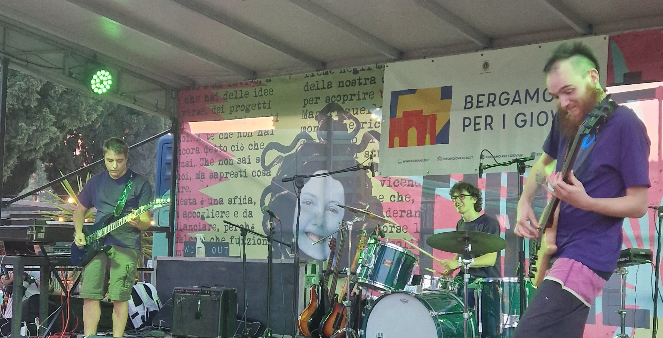 Una band sta suonando durante un concerto su un palco. Da sinistra il bassista, al centro in seconda fila il batterista, a destra il chitarrista.