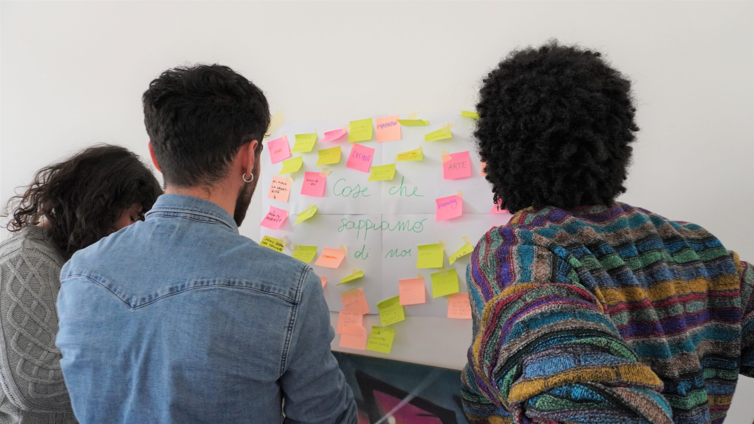 tre persone appendono post-it su un cartellone attaccato a una parete. Sul cartellone c'è scritto: Cose che sappiamo di noi
