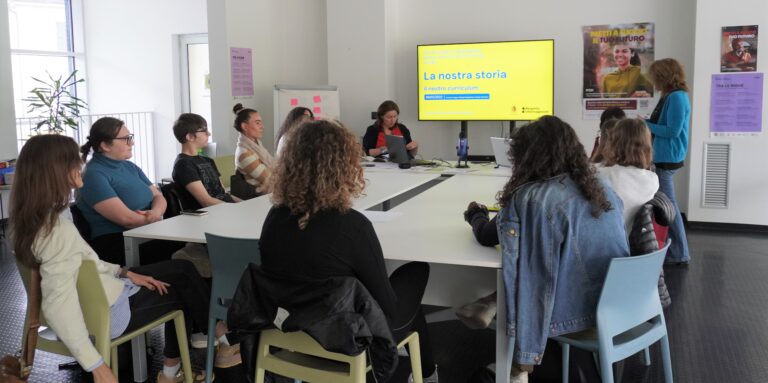 Fotografia di un incontro di gruppo, ci sono diverse persone intorno a un tavolo, una tv che proietta delle slide e una persona in piedi sulla destra