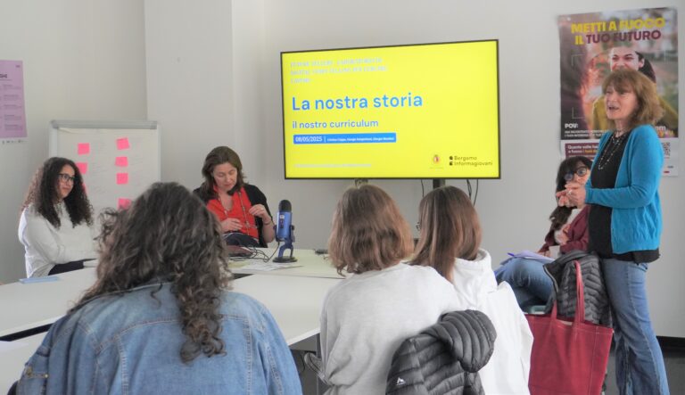 Immagine che ritrae un evento di formazione di gruppo. Una persona sulla destra con maglia blu parla, mentre diverse altre persone la ascoltano e la guardano, sedute intorno a un tavolo
