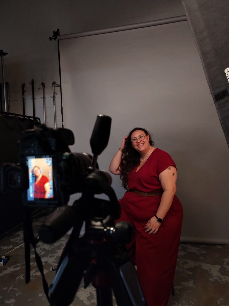 In primo piano a sinistra una macchina fotografica. In secondo piano una persona vestita di rosso su sfondo grigio che viene fotografata