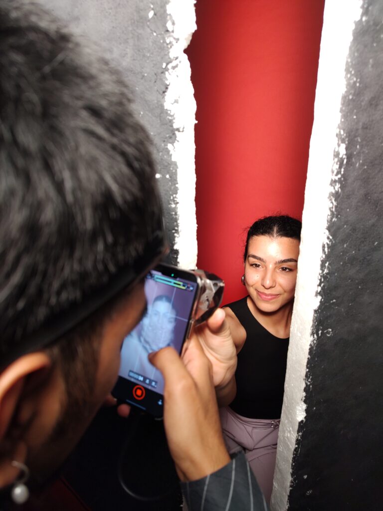 Immagine di backstage fotografico. Una persona fotografa un'altra persona con il telefono. Il viso della persona che viene fotografata sta tra due pannelli e dietro di lei c'è uno sfondo rosso.