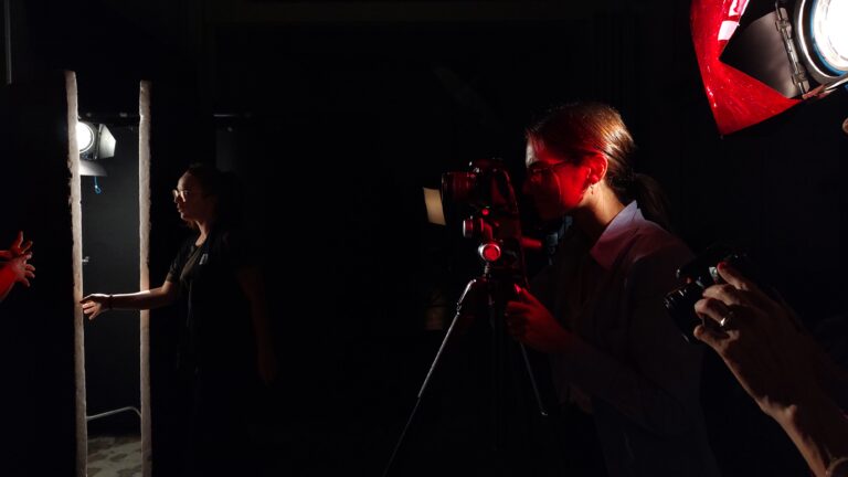 Immagine di un backstage fotografico. Sulla sinistra di chi osserva ci sono due persone che sistemano pannelli accanto a un fascio di luce, sulla destra una luce rossa e persone che fotografano