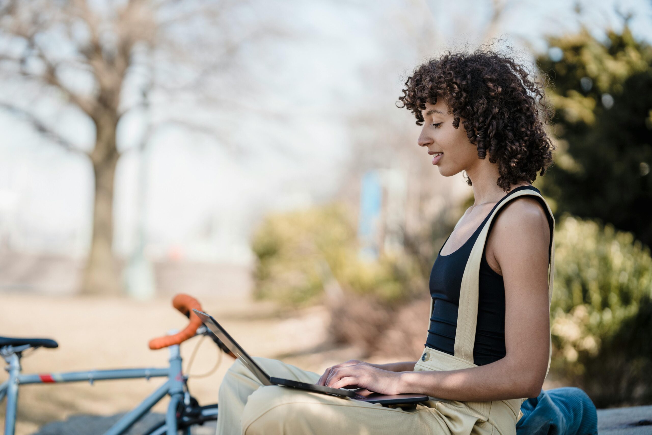 Giovane che studia al pc all'aperto. È seduta, ha le gambe incrociate, una maglietta scura e pantaloni chiari, ha una bici vicino.