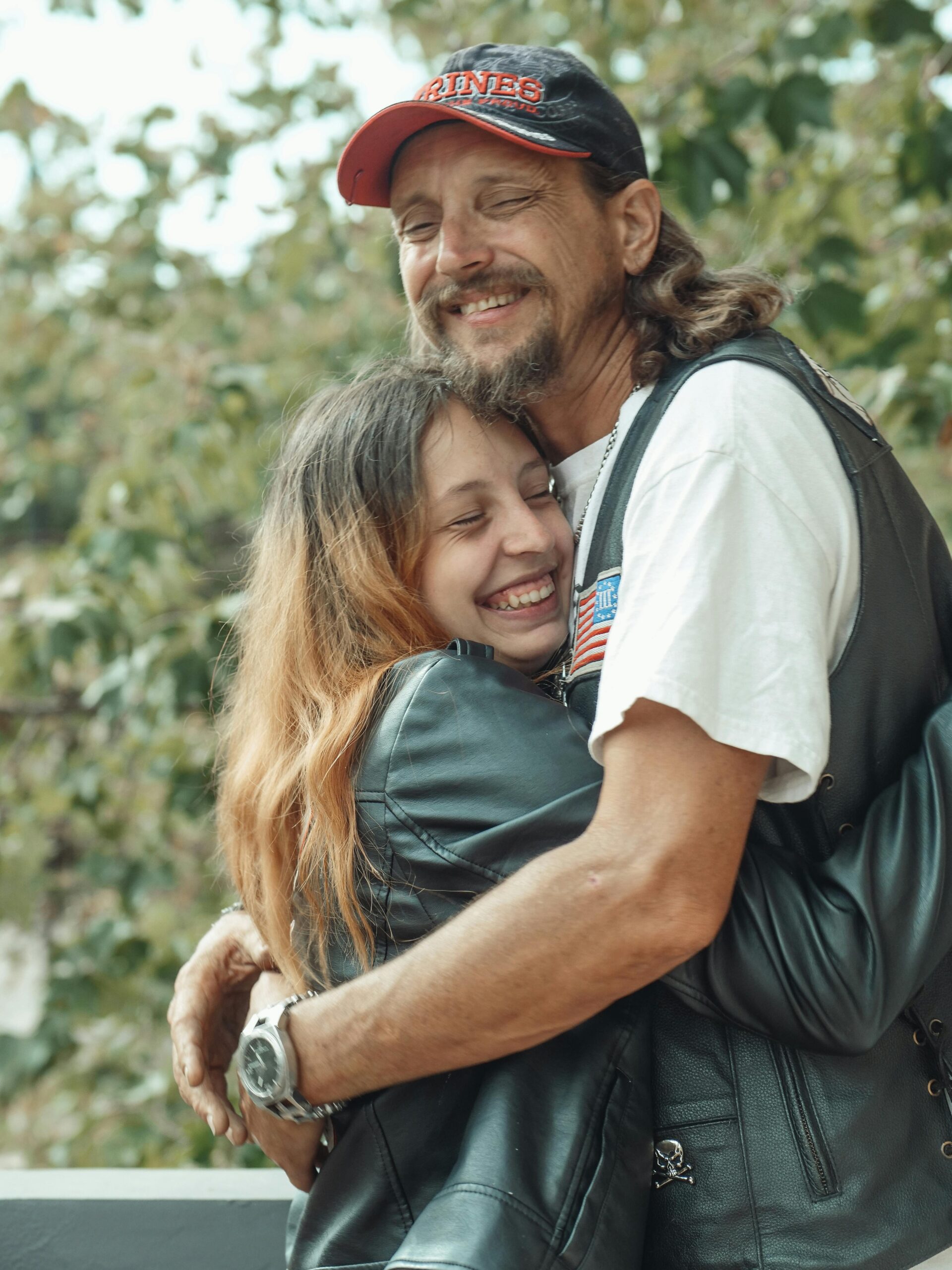 foto di due persone che si abbracciano. Sono una famiglia e il genitore ha i capelli lunghi e un cappello da baseball, la persona più giovane porta una giacca nera. Entrambe le figure sorridono con gli occhi chiusi.