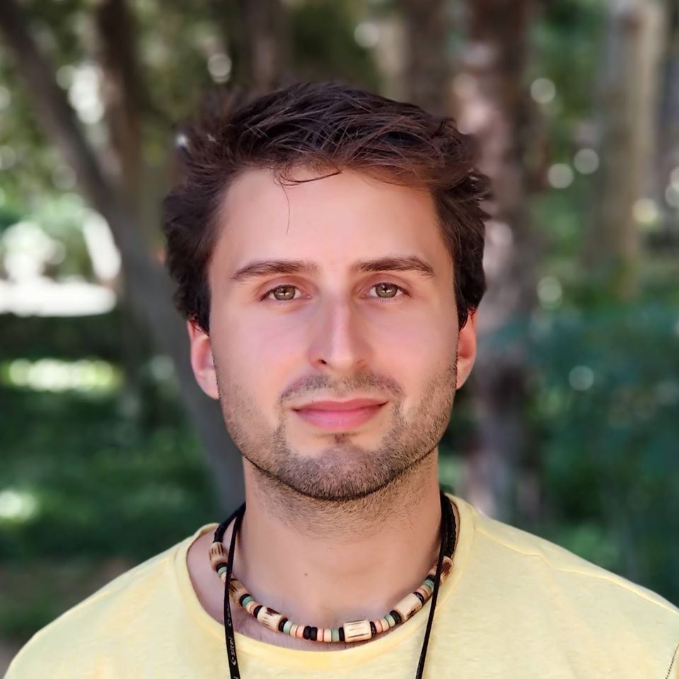 Primo piano di Alvaro, indossa una collana e una maglietta giallina, sullo sfondo ci sono degli alberi.