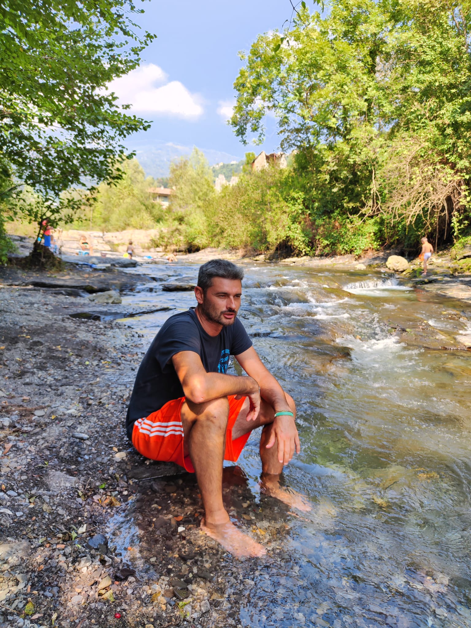 Angelo, con indosso maglietta nera e shorts arancioni. E' seduto su un sasso nel letto di un ruscello.