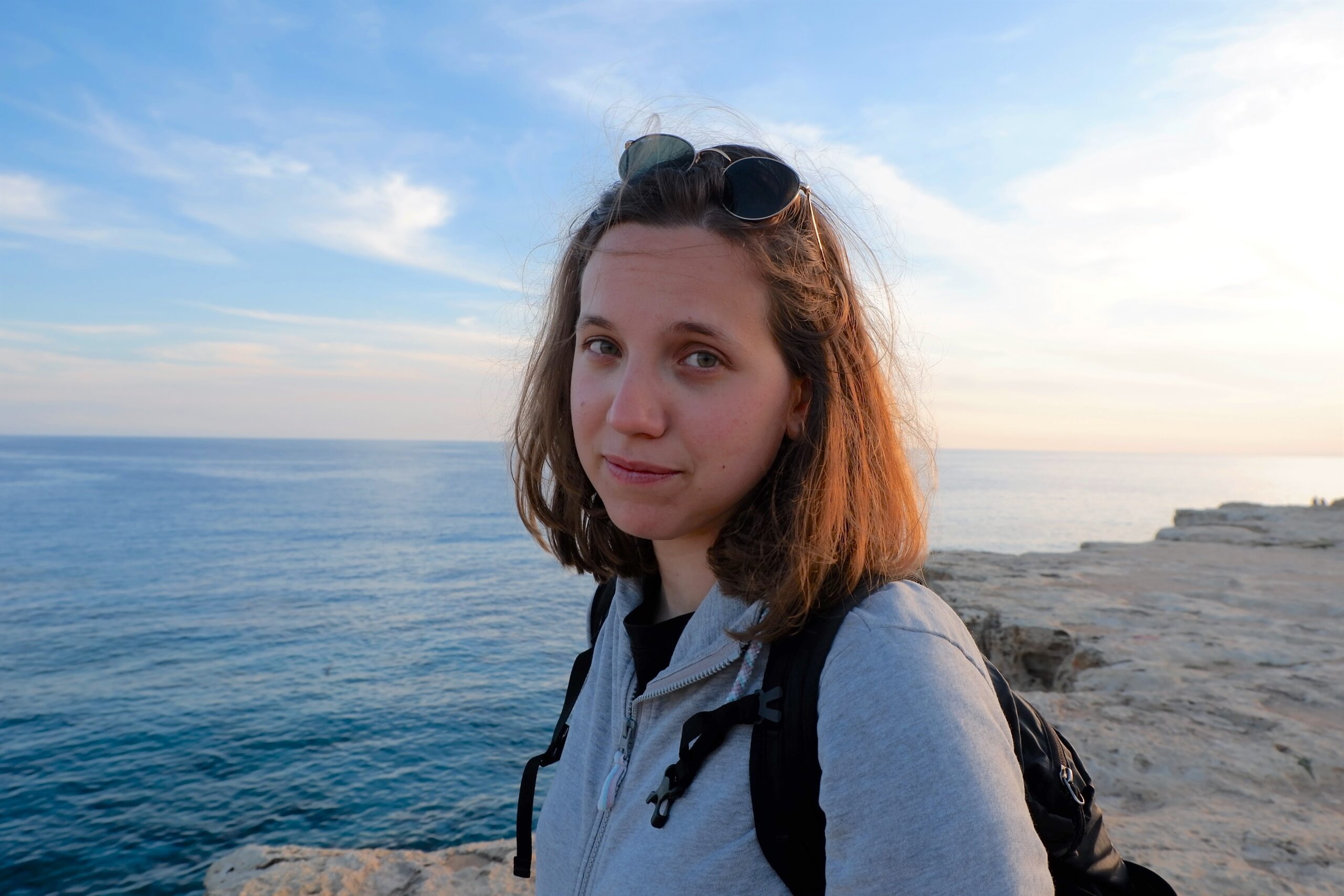 Chiara in riva al mare. Porta gli occhiali da sole appoggiati in testa, indossa una felpa grigia e uno zaino.