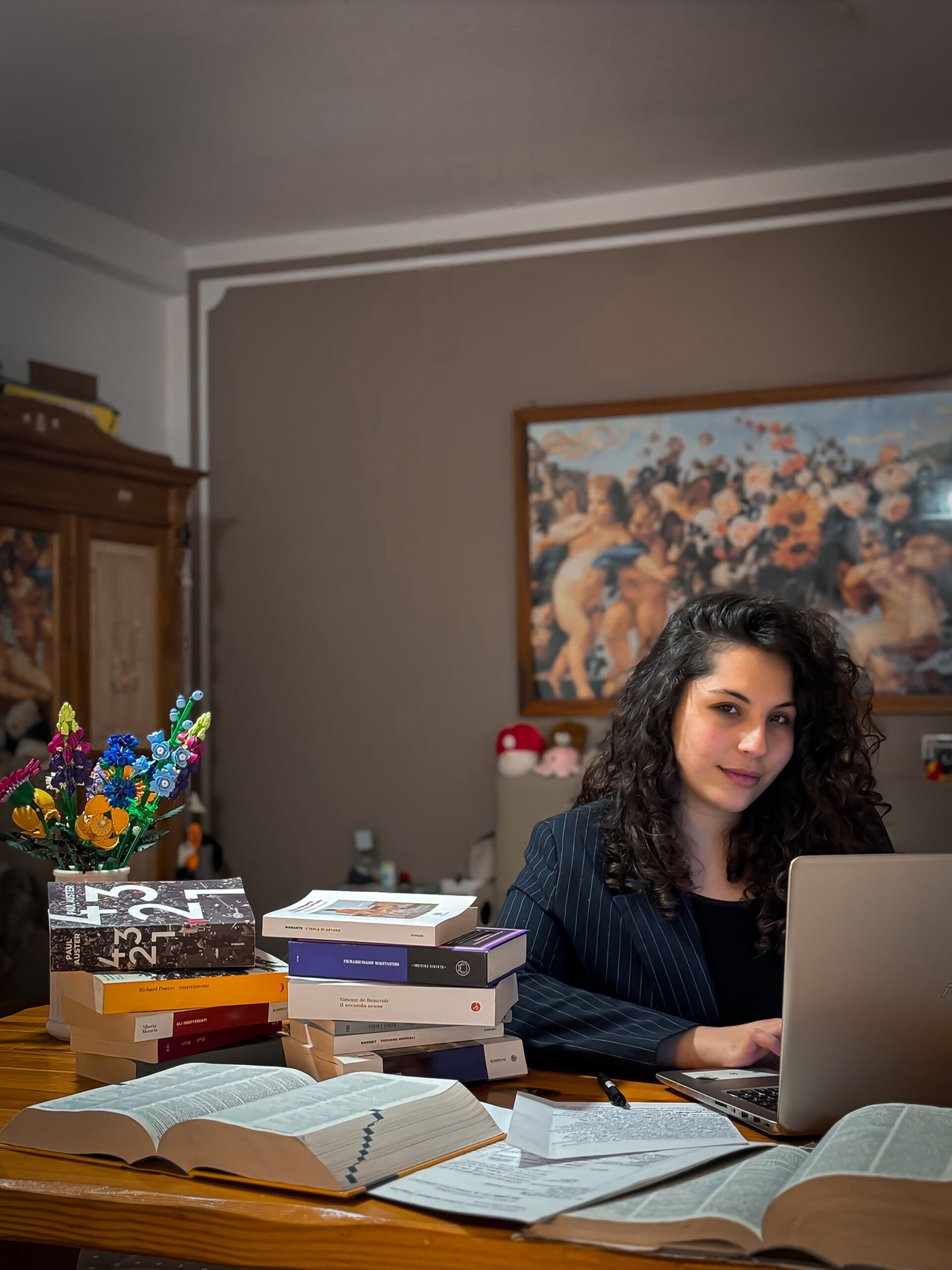 Alessia seduta ad una scrivania piena di libri, sullo sfondo è visibile un quadro.
