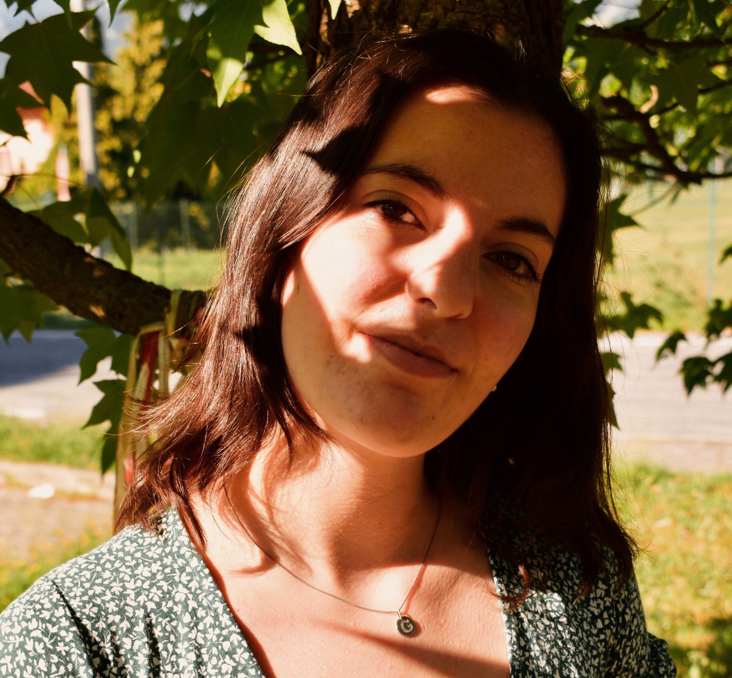 Giulia in primo piano sorride. Indossa un vestito a fiori, una collana con la sua iniziale ed è in piedi sotto un albero