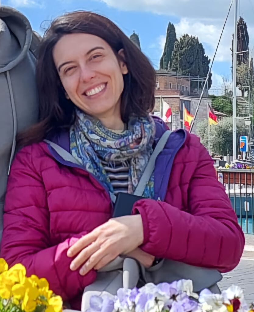 Chiara che sorride con indosso una giacca magenta e una bandana blu, di fronte a lei sono poggiati due vasi di fiori