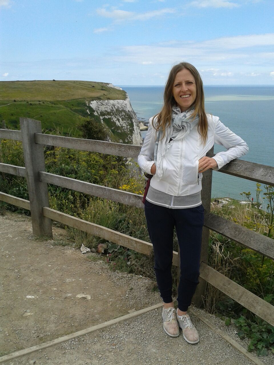 Michela sorridente, indossa un giubbino bianco e pantaloni scuri. Sta in posa appoggiata ad un recinto di legno. Sullo sfondo sono visibili grandi scogliere.