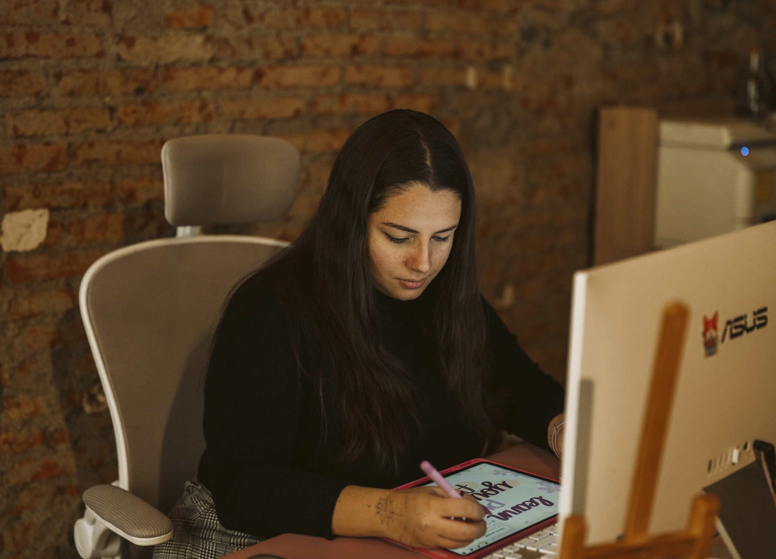 Sara seduta ad una scrivania, lavora con un tablet. Indossa una maglia nera e pantaloni a quadri.