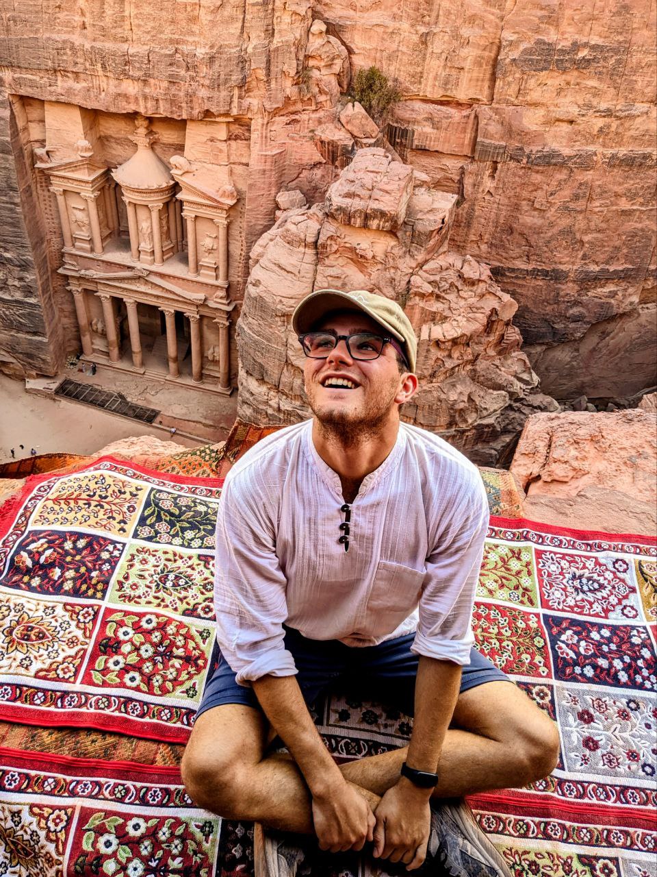 GIovanni seduto su un tappeto colorato di fronte alle rovine di Petra, guarda verso l'alto e indossa una maglietta bianca e un cappello
