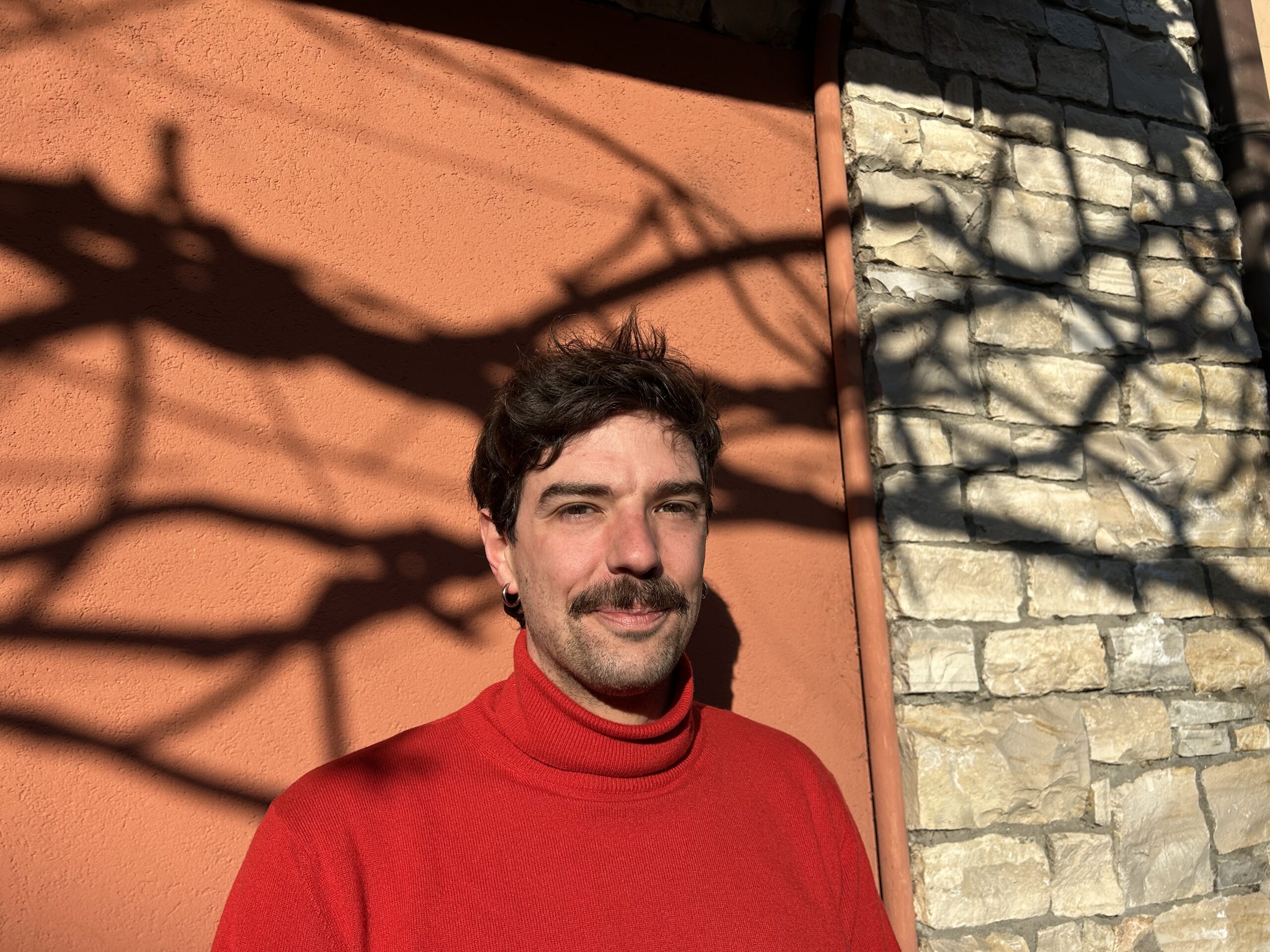 Gregorio, con indosso un maglione rosso, è di fronte a un muro di mattoni.