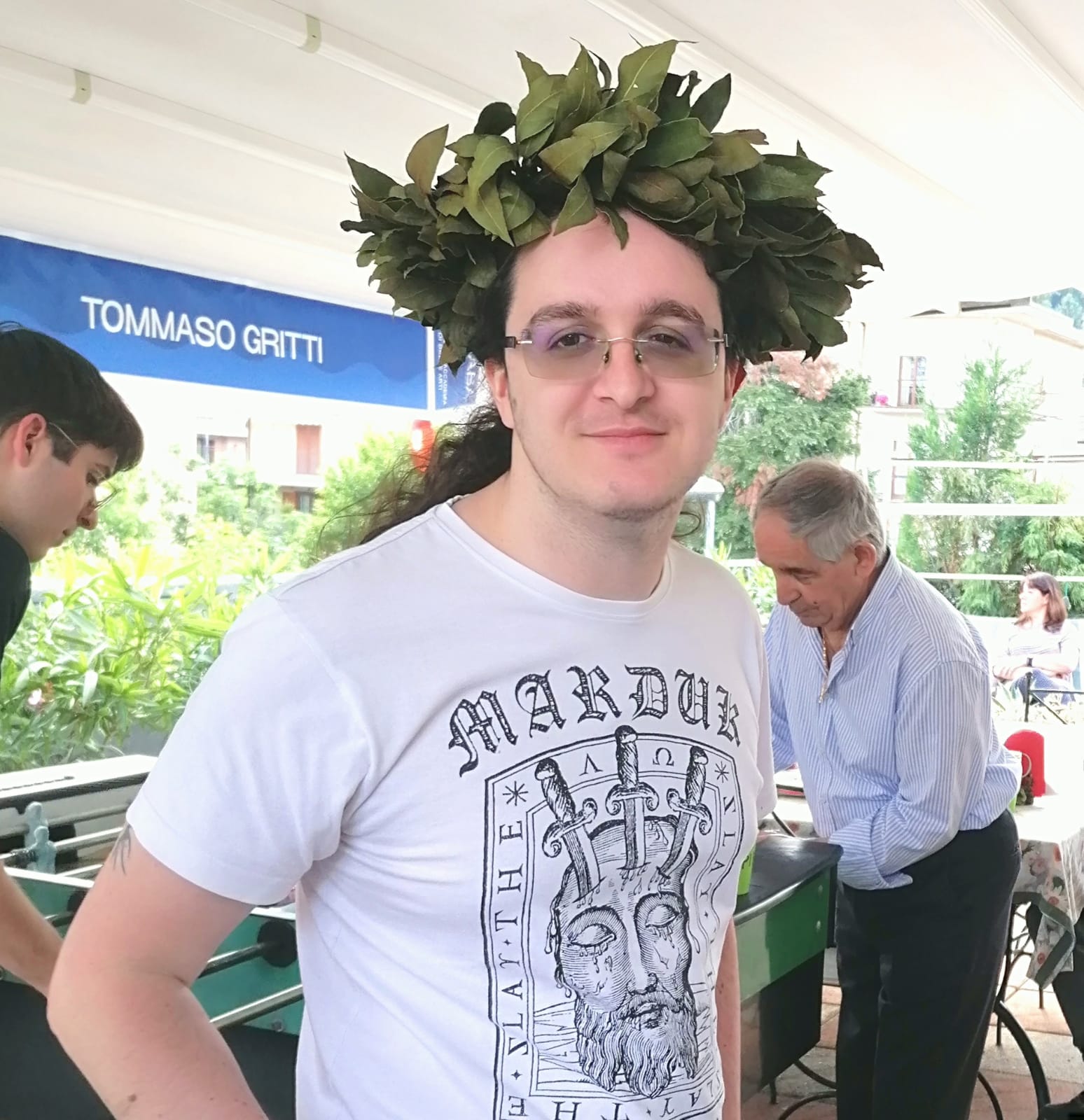 Tommaso con indosso la corona di alloro e una maglia bianca, sorride. dietro di lui delle persone giocano a calcetto.