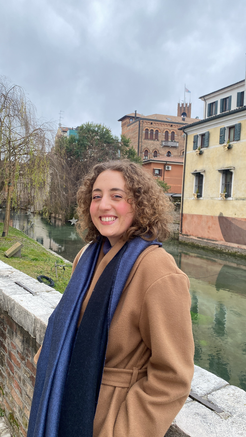 Vanessa sorride mentre è in posa su un ponte. Indossa un cappotto marrone e una sciarpa blu. alle sue spella un canale e degli edifici.