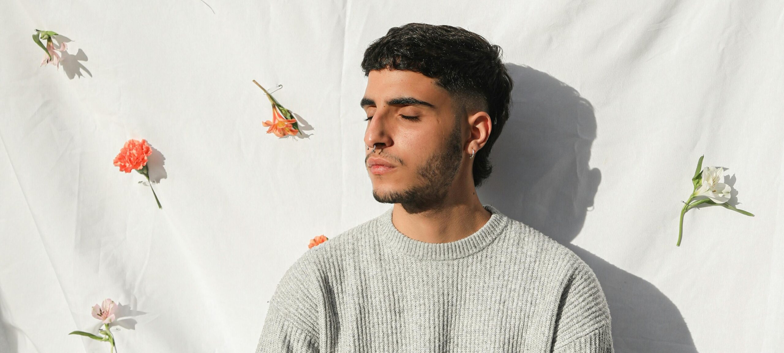 Primo piano di un ragazzo con gli occhi chiusi. Ha i capelli scuri e indossa un maglione grigio. Dietro di lui è steso un lenzuolo bianco a cui sono appesi dei fiori colorati.