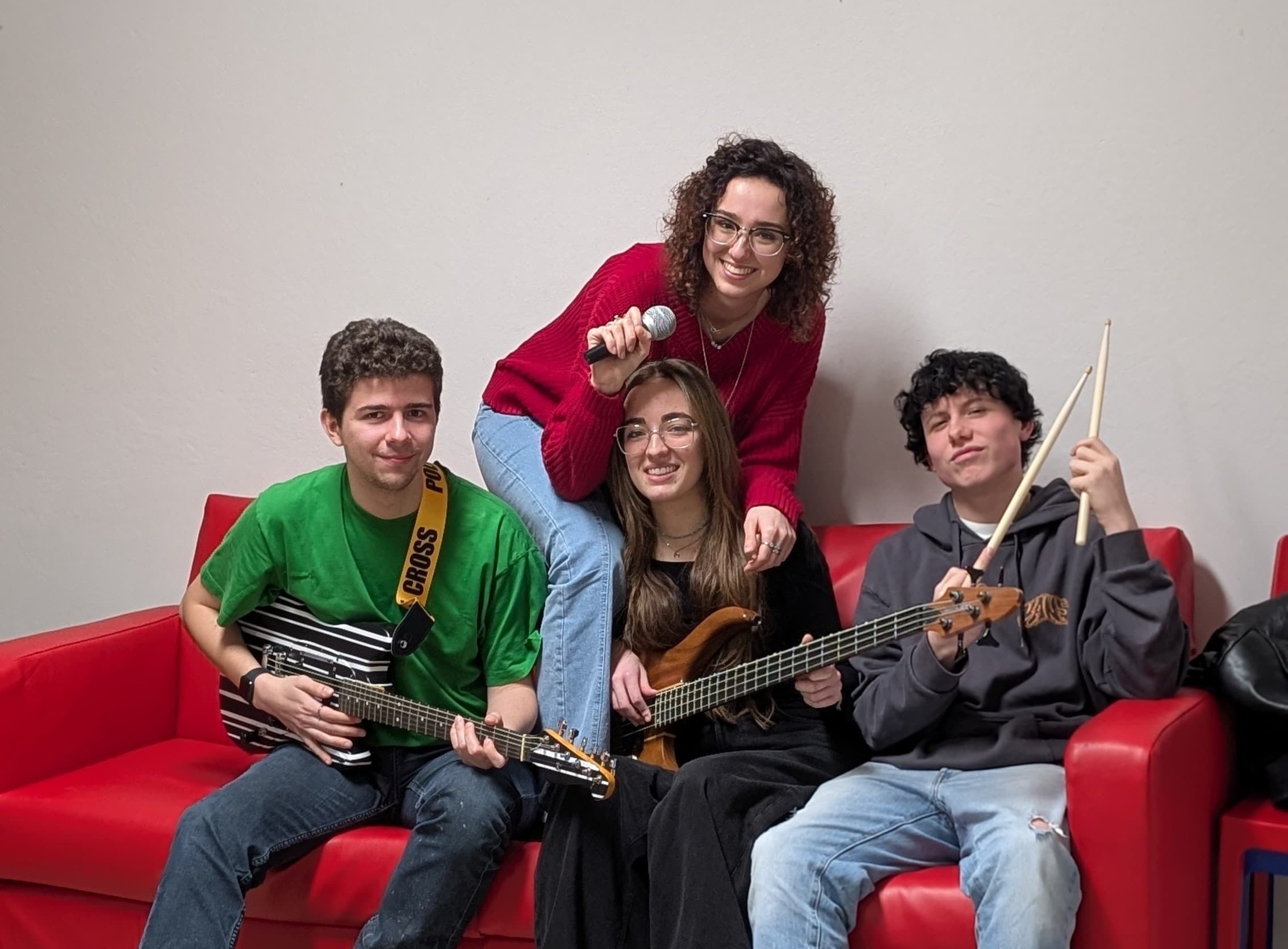 Foto della formazione di una band, seduta su un divano rosso. Da sinistra il chitarrista, la bassista e il batterista. Dietro di loro la cantante.