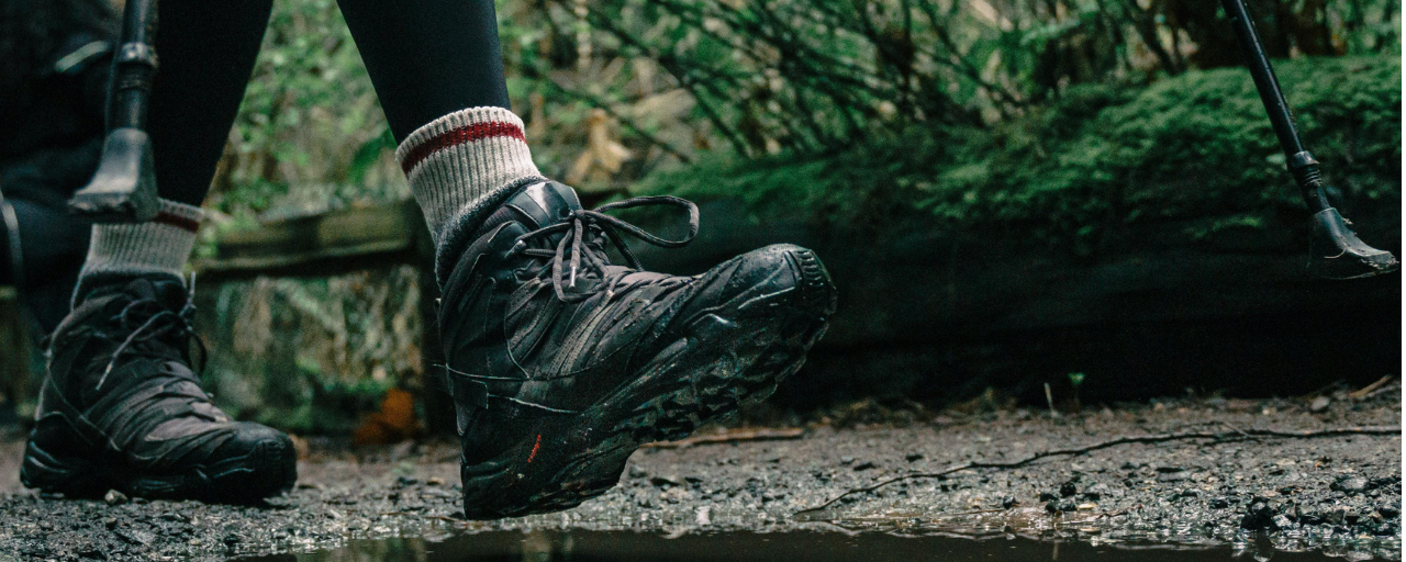 Particolare di una foto: in primo piano due piedi con scarpe da trekking e calzini di spugna sportivi. Lo sfondo è quello di un sentiero in un bosco.