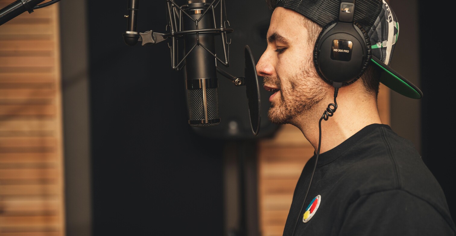 Un ragazzo, fotografato di profilo canta in un microfono in uno studio di registrazione. Indossa grandi cuffie, una felpa nera con un piccolo stemma colorato e un cappello con la visiera portata dietro.