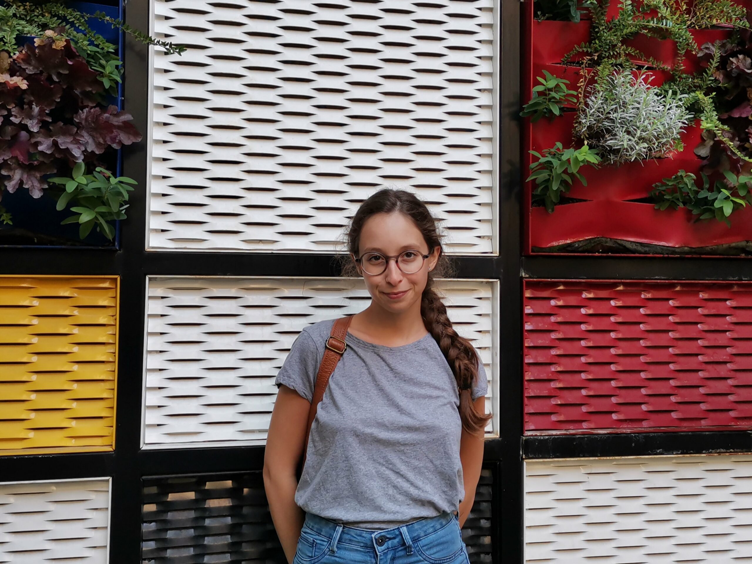 Primo piano di Anna davanti ad un muro colorato. Ha i capelli acconciati in una treccia e indossa una maglia grigia.