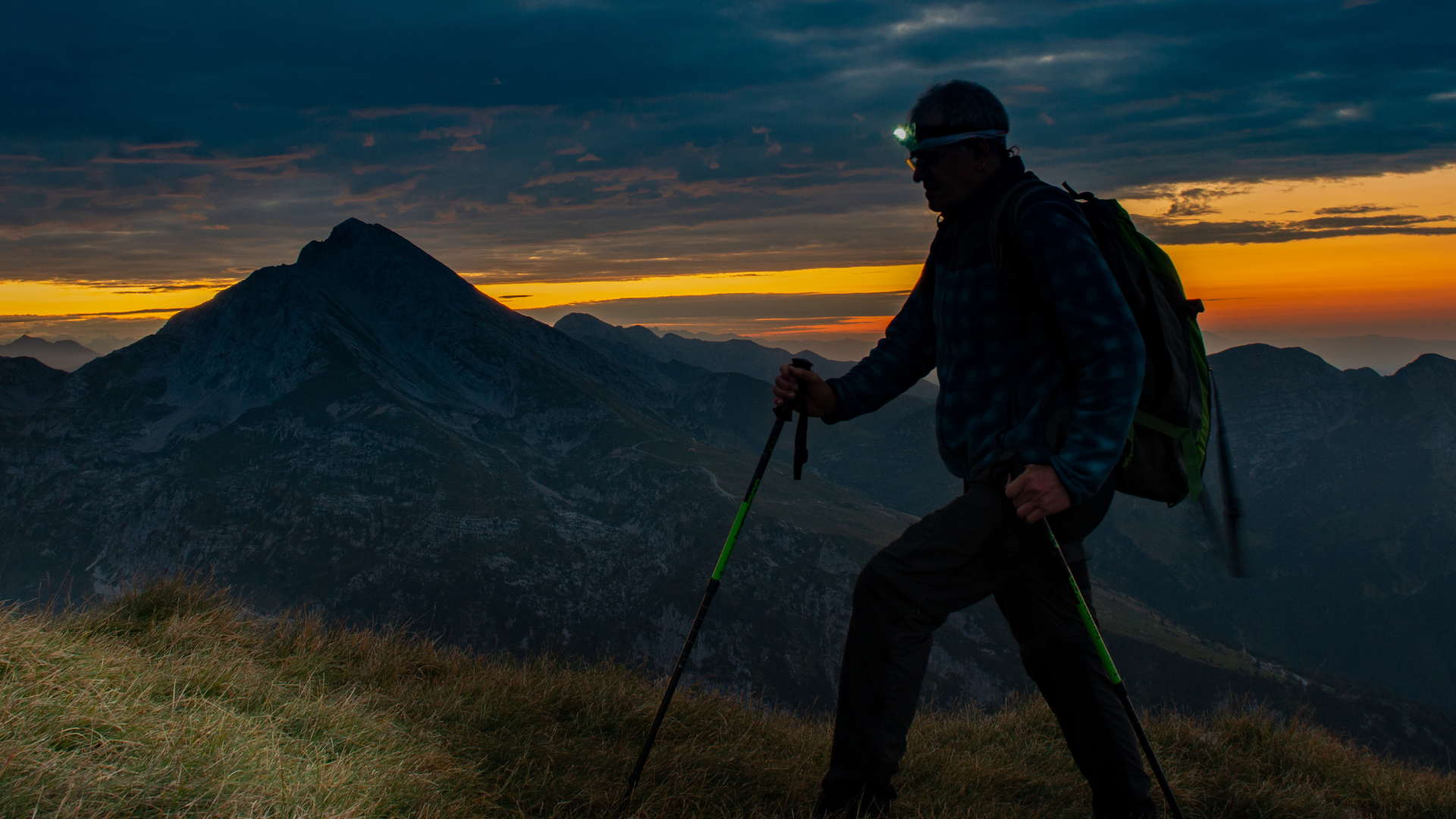 Un escursionista percorre un sentiero di montagna nel buio della notte. Utilizza delle racchette e indossa una luce frontale sulla fronte.